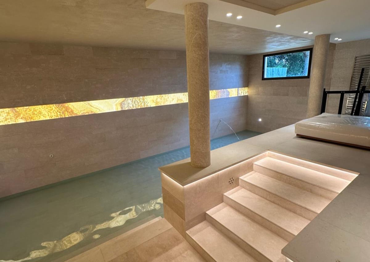 Piscine intérieure habillée en marbre Botticino avec bande décorative en onyx rétroéclairé et escalier en pierre naturelle.
