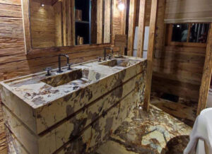 Salle de bains haut de gamme avec double vasque sur mesure en pierre naturelle, murs en vieux bois et ambiance chaleureuse de chalet de prestige.